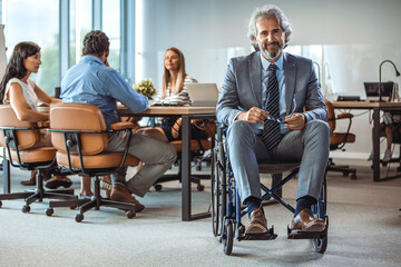 Portrait of mature and cheerful male businessman in a wheelchair holding his smartphone and smiling while sitting at his workplace in modern office. Disabled people