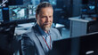 © Gorodenkoff - Portrait of Positive Professional Technical Controller Sitting at His Desk with Multiple Computer Displays Before Him. In the Background His Colleagues Working in System Control and Monitoring Center.