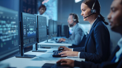 Wall Mural - Happy Beautiful Technical Customer Support Specialist Having a Headset Call while Working on a Computer in a Dark Monitoring and Control Room Filled with Colleagues and Display Screens.