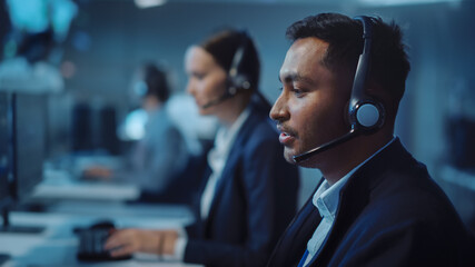 Fototapete - Close Up Portrait of a Joyful Technical Customer Support Specialist Talking on a Headset while Working on a Computer in a Call Center Control Room Filled with Computer Display Screens and Data Servers