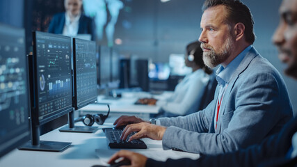 Fototapete - Portrait of Serious Professional Technical Controller Sitting at His Desk with Multiple Computer Displays Before Him. In the Background His Colleagues Working in System Control and Monitoring Center.