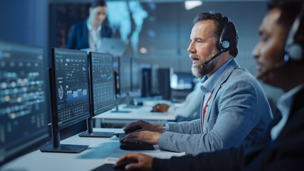 Fototapete - Bearded Happy Senior Technical Support Specialist is Talking on a Headset while Working on a Computer in a Dark Monitoring and Control Room Filled with Computer Display Screens and Data Servers.