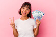 © Asier - Young mixed race woman holding bills isolated on pink background joyful and carefree showing a peace symbol with fingers.