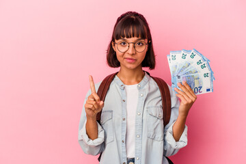 Wall Mural - Young mixed race student woman holding bills isolated on pink background showing number one with finger.