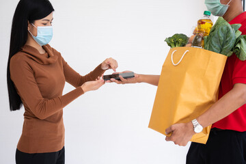 Asian delivery man holding holding a bag of fresh food for giving to customers and holding smartphone for Receive payments at home. Concept of express grocery service and new lifestyle