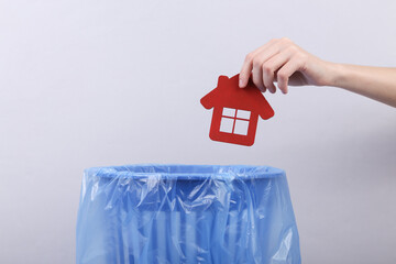 Hand throws house figure into trash bin with package on gray background