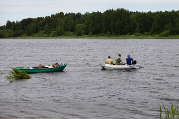 Wall Mural - Three caucasian fishers without lifejackets floats on modern inflatable motor boat with loaded rowing boat on rope, fishing recreation on the Russian river at summer day