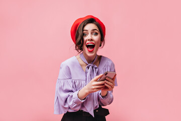 Canvas Print - Emotional woman in red beret and stylish shirt in joyful surprise looks at camera and holds white smartphone on pink background