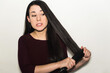 © AliciaFdez - Confused young woman straightening her long black shiny hair using a flat iron isolated on a white background