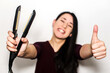 © AliciaFdez - Smiling young woman holding a flat iron in one hand and doing the thumbs up sign with the other. Hair straightener
