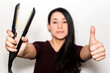 © AliciaFdez - Pretty european young woman holding a flat iron in one hand and doing the thumbs up sign with the other. Hair straightener