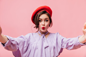 Wall Mural - Girl in lilac blouse and red beret blows kiss and takes selfie on pink background