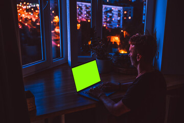 Wall Mural - a laptop with an empty green screen on a wooden table at night with a view of the city with colored 