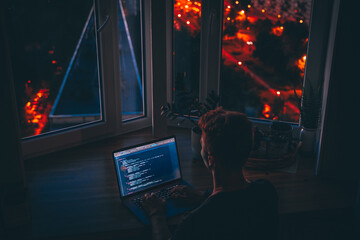 Wall Mural - a young man programmer coding on a laptop in the dark with a view of the lights of the night city, c