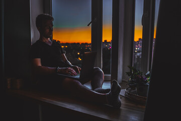 Wall Mural - A young programmer is coding on a laptop in the dark sitting on a window with a view of the lights of the night city, colored lighting in the room, home decor