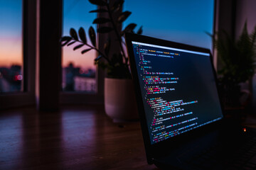 Wall Mural - the code is on a laptop on a wooden table in front of the window in the dark with a view of the ligh