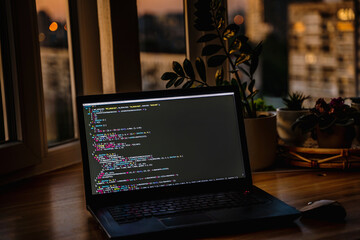 Wall Mural - the code is on a laptop on a wooden table in front of the window in the dark with a view of the ligh
