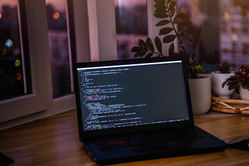 Wall Mural - the code is on a laptop on a wooden table in front of the window in the dark with a view of the ligh