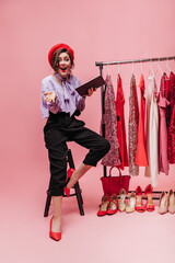 Wall Mural - Brunette girl in red beret laughs sitting on chair with folder in her hands. Lady with red lipstick posing on pink background with stand with shiny dresses