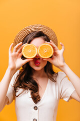 Wall Mural - Cheerful woman with wavy hair covers her eyes with oranges. Lady in boater and shirt posing on isolated background