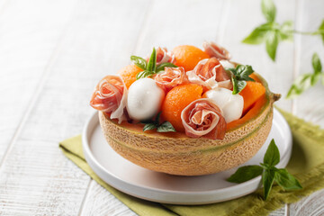 Wall Mural - Italian summer salad. Prosciutto, mozzarella cheese and melon balls in melon basket, decorated with basil leaves. White background, copy space. Selective focus.