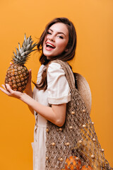 Poster - Attractive long-haired woman in white dress posing with knitted shopping bag and holding pineapple