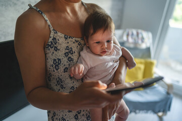 Adult woman mother holding three months old baby while using looking mobile phone for internet browsing or social network texting in room at home domestic life motherhood and parenting busy concept