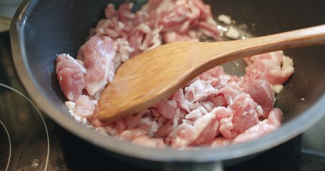 Close up wooden spoon stirring sizzling cooking stir frying fresh raw meat slices pork in hot pat on electric stove