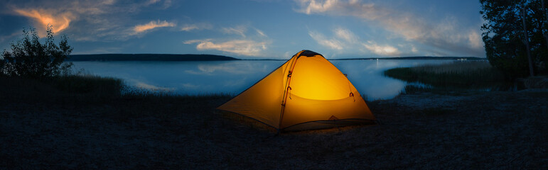 Wall Mural - orange tourist tent illuminated from inside on shore of lake under sunset sky