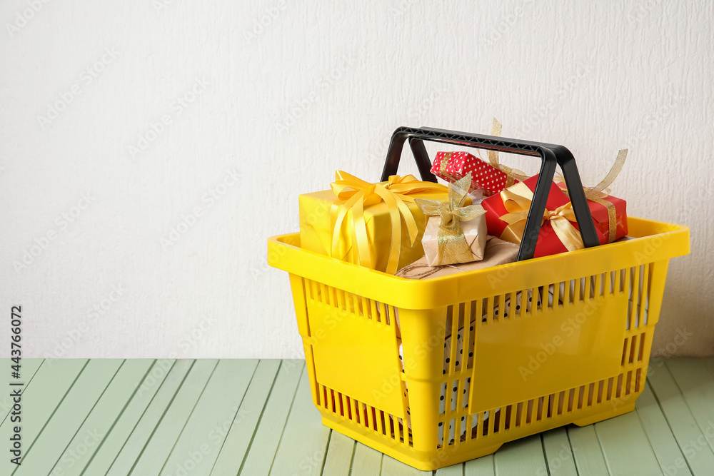 Shopping basket with gift boxes on table near white wall