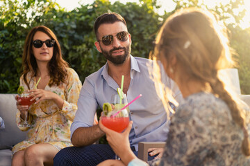 Group of young people talking together having fun while drinking soft drinks and find themselves together sitting on a sofa outdoors in the garden.