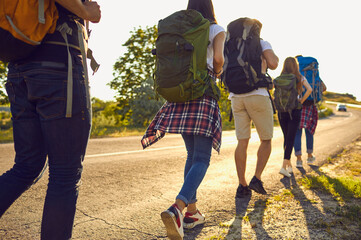 Wall Mural - Group of young travelers hikers hiking and hitch-hiking on road with backpacks together during summer vacations on sunny clear day, rear view. Hiking and traveling together concept