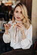 © Look! - Blonde young woman in white blouse, dark pants and pearl necklace spreads paste on bruschetta and poses in restaurant. Blurred effect.
