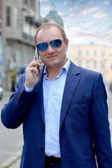 Young caucasian man in a blue business suit and sunglasses holds a smartphone for business negotiations. Business, fast pace of life concept. Outside the office in the historic city center. Europe