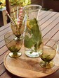 Three glasses and bottle fill with water, lemon slice and lemon balm. One glass filled with paper straws. All standing on round wooden tray outside. View at an angle. 