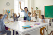 © Studio Romantic - Happy clever enthusiastic school children and teacher raising hands. Confident preschoolers or first grade students sitting around big classroom table, learning letters, reading books and having fun