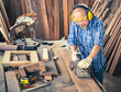 © panyajampatong - Happy senior carpenter using an electric planer with a wooden plank in the carpentry workshop. Manufacture of wood products..