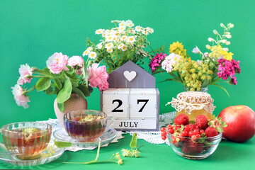 Wall Mural -  Calendar for July 27 : the name of the month of July in English, cubes with the number 27, bouquets of wild flowers, jam, fruit, cups of tea, white napkins, green background