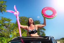 Horizontal Shot Of Attractive Female With African Braids In A Black Swimsuit And An Inflatable Ring And A Hare In Her Hands, Looking Out Of The Car Hatch And Smiling. Middle Aged Woman On Sunny Beach