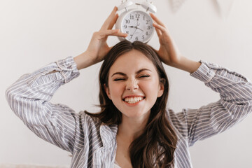 Wall Mural - Woman in pajamas makes nice face and keeps alarm clock on her head. Portrait of brunette girl laughing. Teen in shirt posing on white background