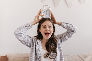 Wall Mural - Green-eyed girl enthusiastically looking at camera and holding alarm clock on her head. Dark haired woman in blue shirt and tee happily posing on white backgroung