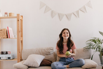 Wall Mural - Joyful lady in jeans and t-shirt laughs, sits on couch and holds apple. Healthy girl in striped tee holing fruits and posing at home