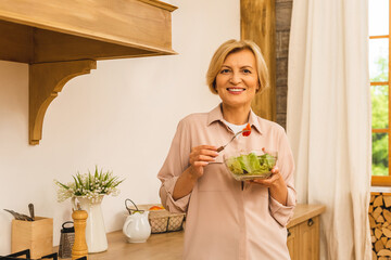 Modern senior aged mature woman eating fresh green salad and vegetables in kitchen, smiling happy. Helthy lifestyle concept.