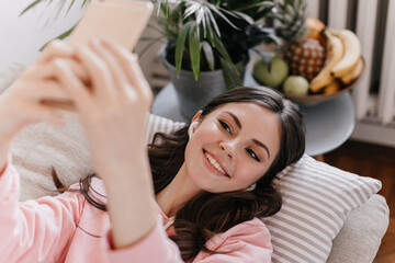 Portrait of charming girl lying on pillow in living room and taking selfie. Brunette teen smiling. Curly woman in pink home outfit holds cellphone