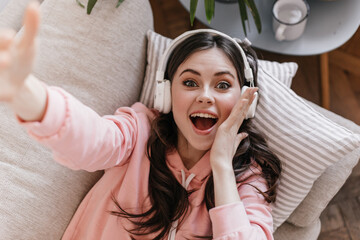 Wall Mural - Emotional girl in white headphones takes selfie and looks at camera in surprise, lying on couch. Brunette woman taking selfie in cozy home atmosphere