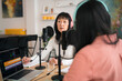 © Studio Marmellata/Stocksy - Asian woman speaking to microphone during podcast recording with colleague