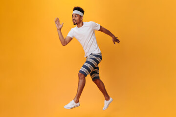 Wall Mural - Dark-skinned guy in shorts and T-shirt runs on orange background. Sportsman has workout on isolated. Portrait of dark-skinned teen