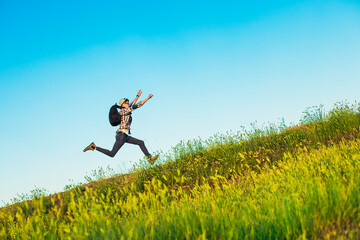 Wall Mural - Happy young tourist with a backpack, happy jumping man with hands up against a blue sky background, climbs to the top of a hill or mountain