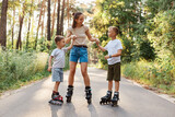 Outdoor shot of happy family having fun and roller skating together in summer park, mommy holding kids hands, being glad to spend weekend together, active pastime.