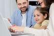 © LIGHTFIELD STUDIOS - blurred woman pointing at laptop near daughter and smiling husband